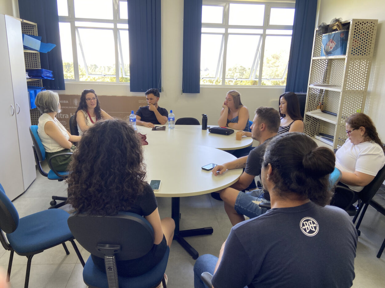 A foto mostra a equipe da Universidade Estadual de Ponta Grossa reunida em uma sala no campus. Todos estão sentados em um grande círculo ao redor de duas mesas. Ao fundo duas grandes janelas iluminam o ambiente. Uma articuladora da Rede tem a palavra, e os demais ouvem atentamente, parecendo reflexivos.