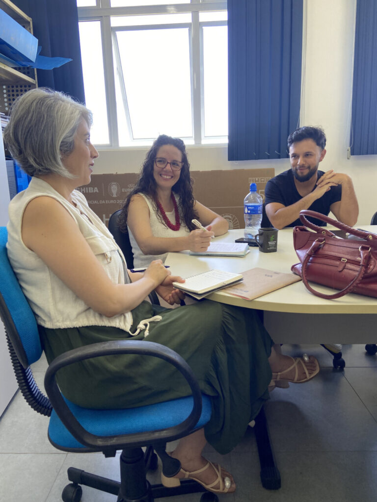 Três membros da equipe dos clubes da UEPG estão sentados ao redor de uma mesa em uma sala de reunião. À esquerda, uma mulher de cabelo curto grisalho segura um caderno. No centro, uma mulher de cabelo cacheado e óculos sorri, com uma caneta na mão. À direita, um homem de barba curta escuta atentamente. Sobre a mesa há cadernos, uma garrafa de água e uma bolsa.