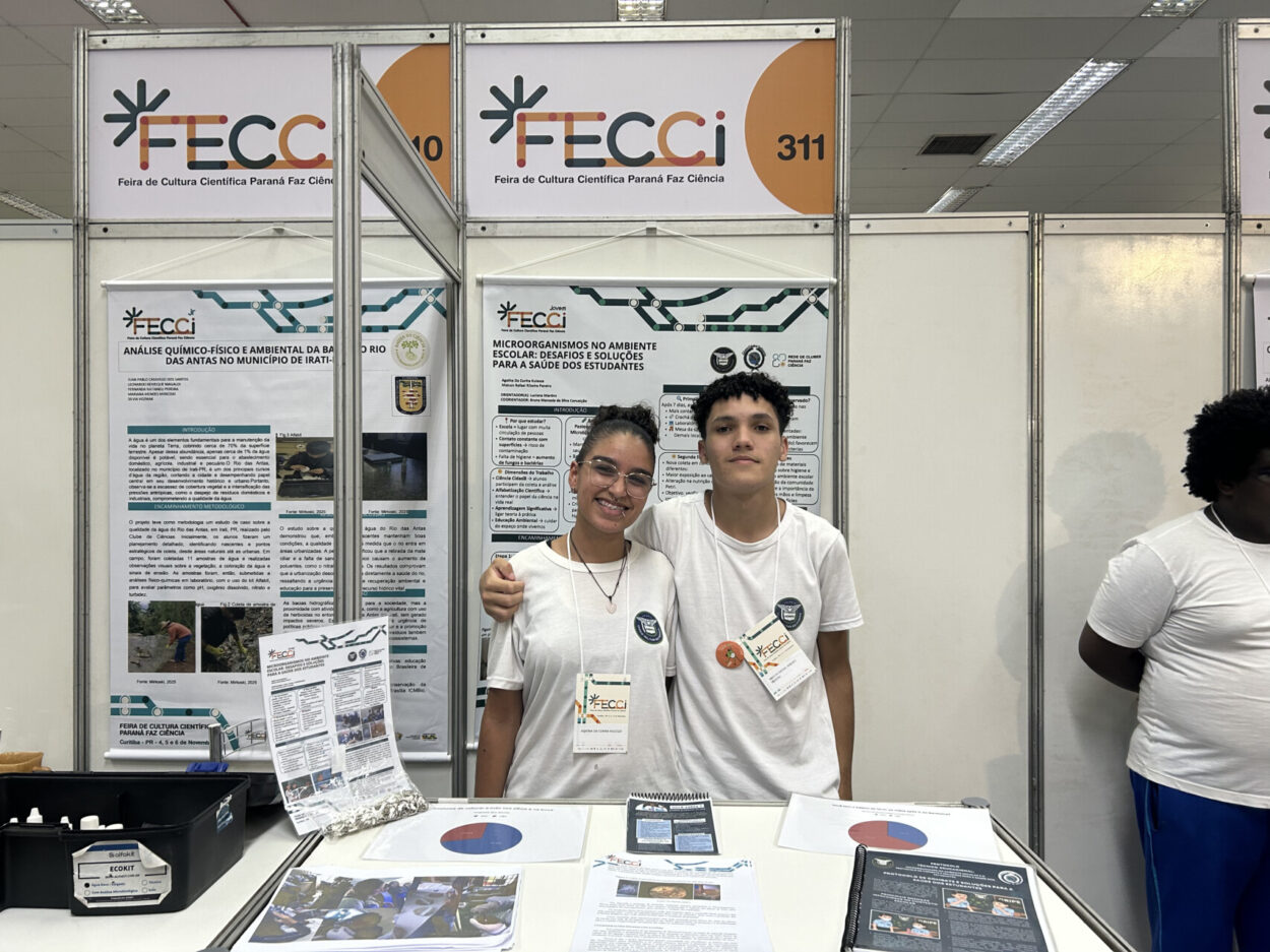 Dois estudantes, usando camisetas brancas e crachás, posam abraçados em um estande da FECCI, com pôsteres científicos ao fundo e materiais de pesquisa sobre a mesa.