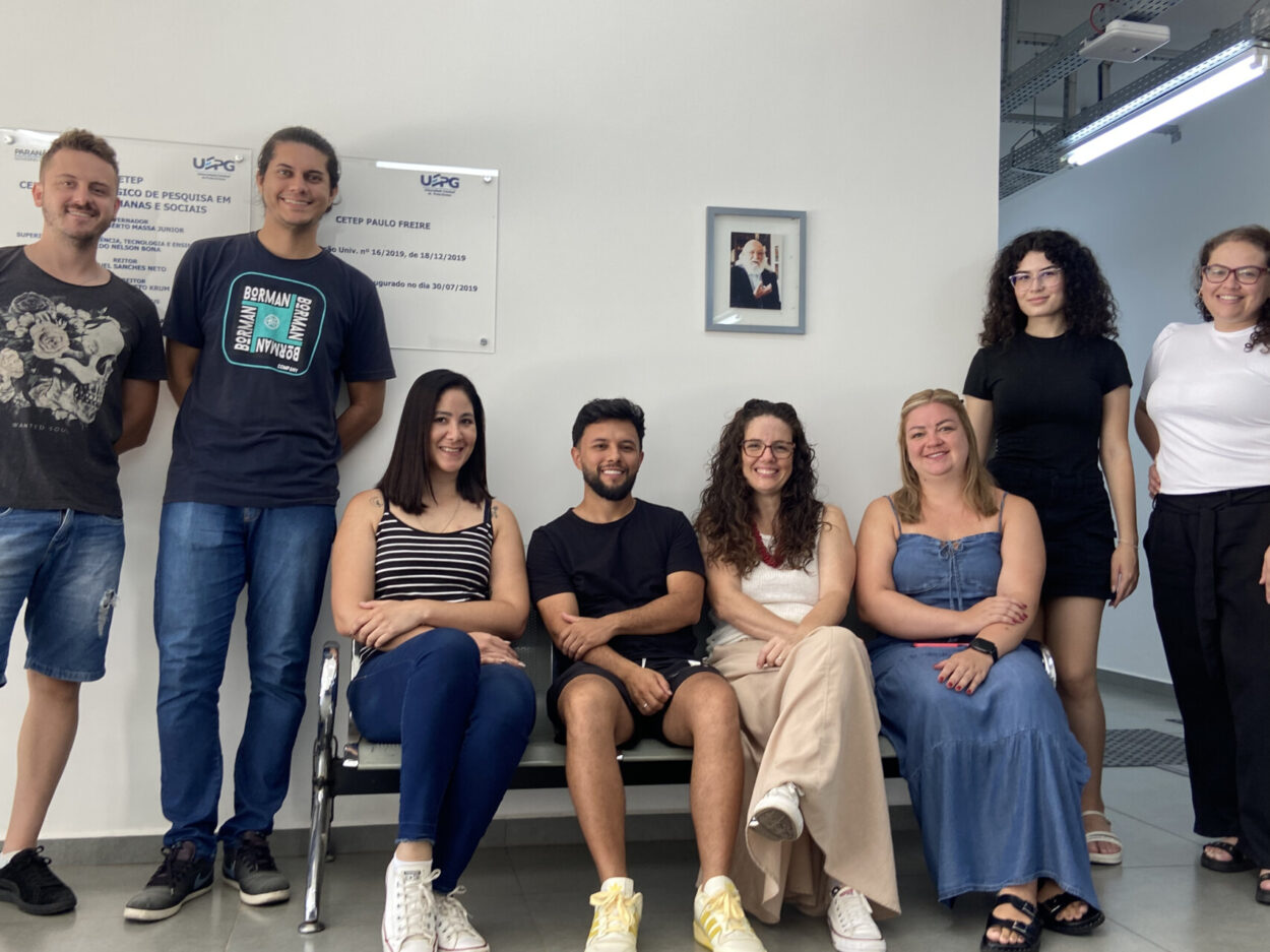 Dez membros da equipe dos clubes da UEPG posam para uma foto em um ambiente interno. Algumas estão sentadas em um banco e outras em pé atrás. Todos olham para a câmera e sorriem. Ao fundo, há uma parede clara com placas institucionais e um retrato de Paulo Freire.