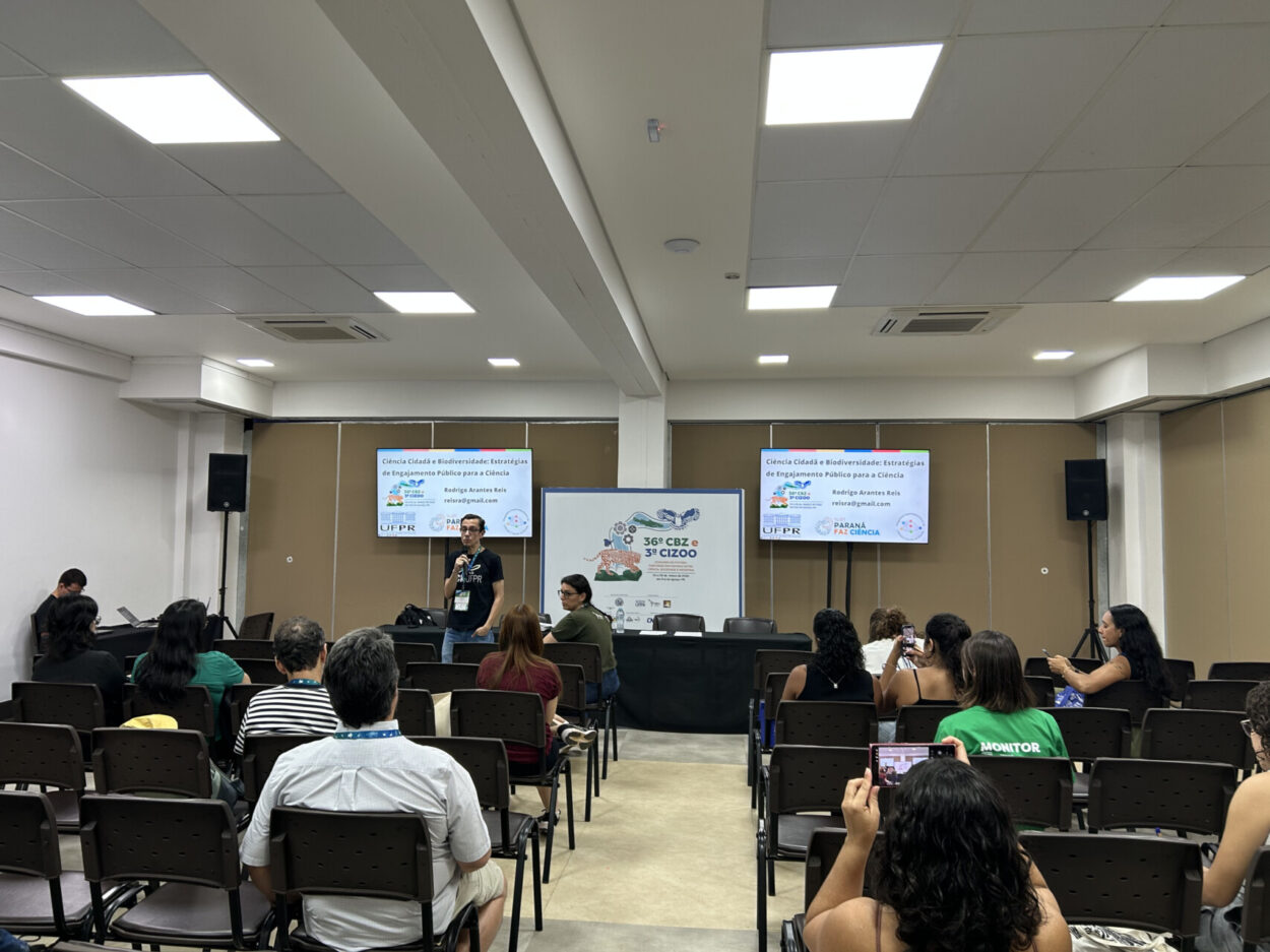 O professor Rodrigo Arantes Reis apresenta a oficina “Ciência Cidadã e Biodiversidade: Estratégias de Engajamento Público para a Ciência” durante o 36º Congresso Brasileiro de Zoologia (CBZ) e a 3ª Conferência da Zoologia na Indústria (CIZOO). Na sala de conferências, participantes estão sentados acompanhando a apresentação enquanto dois telões exibem o título da atividade e logos institucionais, incluindo o Paraná Faz Ciência e a Universidade Federal do Paraná. O ambiente é estruturado para atividades acadêmicas e troca de conhecimentos entre pesquisadores e estudantes.