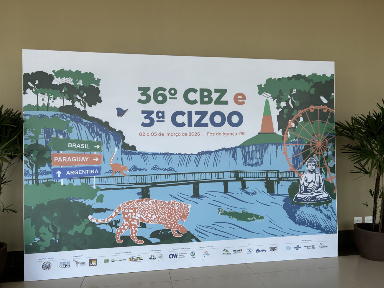 A imagem mostra um grande painel decorativo montado em um espaço interno, utilizado como fundo para fotos em um evento. No centro do painel está escrito “36º CBZ e 3ª CIZOO”, com as datas 02 a 05 de março de 2026, em Foz do Iguaçu. A arte do painel apresenta uma ilustração colorida inspirada na natureza e nos pontos turísticos da região.