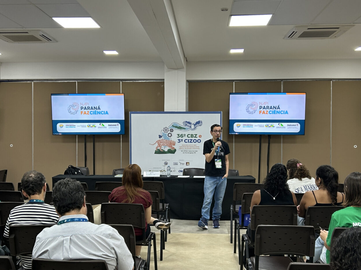 O professor Rodrigo Arantes Reis apresentou uma atividade para participantes durante o 36º Congresso Brasileiro de Zoologia (CBZ) e a 3ª Conferência da Zoologia na Indústria (CIZOO). Ele está em pé, segurando um microfone e falando ao público sentado em cadeiras voltadas para o palco. Ao fundo, duas telas exibem a identidade visual do NAPI Paraná Faz Ciência, enquanto um painel central mostra a marca do congresso. O ambiente é uma sala de conferências com iluminação clara e estrutura para apresentações científicas.