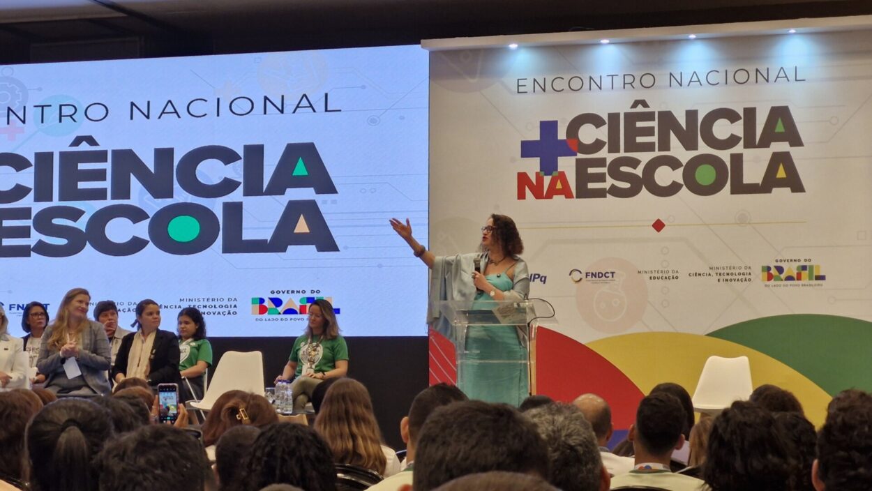 Nesta imagem, vemos um momento formal do Encontro Nacional + Ciência na Escola acontecendo em um auditório. No centro, uma mulher está no púlpito, segurando um microfone e fazendo um gesto com a mão, como se estivesse explicando ou destacando um ponto importante. Ao fundo, há um grande telão com o nome do evento “+ Ciência na Escola” e logotipos de instituições como ministérios e órgãos de ciência e tecnologia. À esquerda do palco, outras pessoas estão sentadas em cadeiras, acompanhando a fala. Na frente, aparece parte do público, que assiste atentamente à apresentação.
