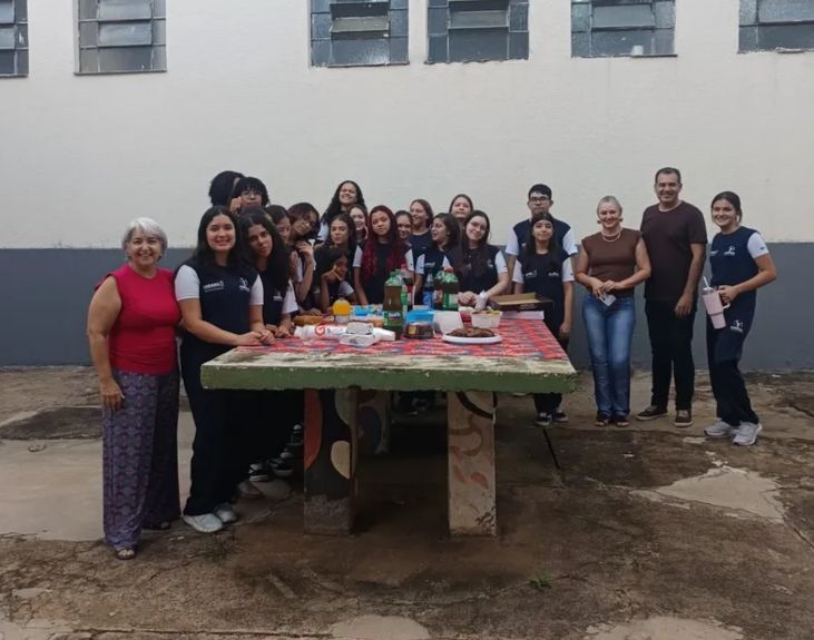 A imagem mostra um grupo de cerca de quinze pessoas, entre adolescentes e adultos, está reunido ao ar livre em frente a uma parede clara com janelas altas. Eles se organizam atrás e ao lado de uma mesa grande de concreto, coberta com uma toalha vermelha estampada. Sobre a mesa há alimentos e bebidas, como garrafas de refrigerante, pratos e recipientes, sugerindo um lanche coletivo ou confraternização. A maioria veste uniforme escolar azul-escuro com detalhes claros. Duas mulheres adultas estão nas laterais do grupo, uma usando blusa vermelha e outra marrom. Todos sorriem ou olham para a câmera, transmitindo um clima de união e celebração.