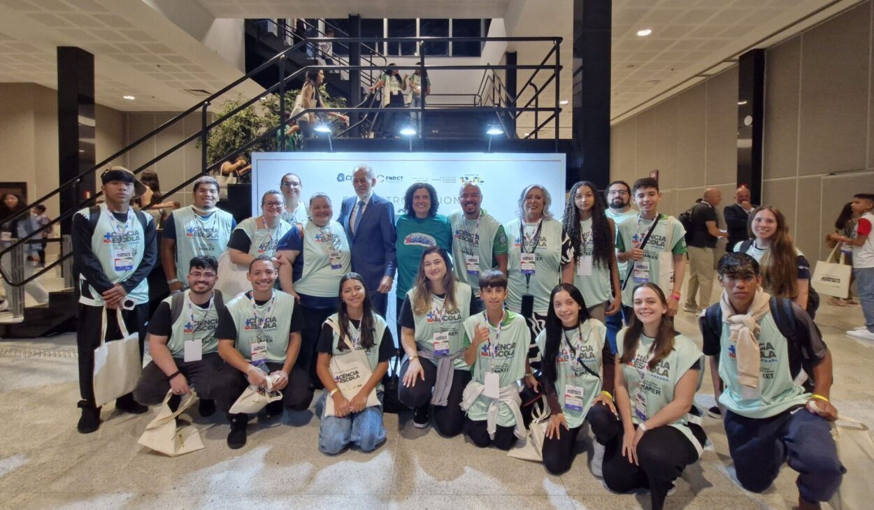 A imagem mostra um grupo grande de estudantes e alguns adultos reunidos para uma foto em um espaço interno. Quase todos usam camisetas verdes do projeto Ciência na Escola e crachás de identificação, indicando participação em um evento. Muitos estão sorrindo e alguns seguram sacolas, provavelmente brindes do encontro. Ao fundo, há uma estrutura com escadas e um painel iluminado com logotipos institucionais. A imagem transmite união, participação e orgulho da equipe. Há ainda a presença de duas autoridades no meio da foto: Juana Nunes e Inácio Arruda.