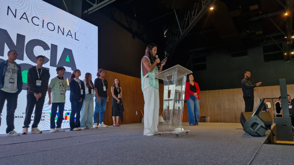 A imagem mostra um palco de evento, em um espaço grande e fechado, com iluminação voltada para a apresentação. No centro, uma jovem está em pé atrás de um púlpito transparente, segurando um microfone e lendo algo, possivelmente um discurso ou apresentação. Ela veste roupas claras e parece concentrada no que está dizendo. À esquerda do palco, há um grupo de cerca de oito pessoas, jovens e adultos, alinhados lado a lado, todos usando crachás pendurados no pescoço, sugerindo que participam do evento como equipe, palestrantes ou competidores.