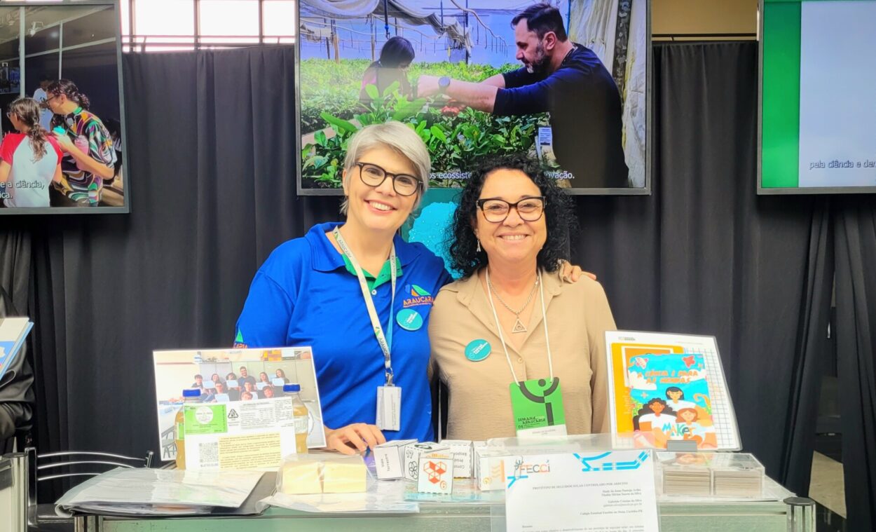 Duas mulheres estão em pé lado a lado, sorrindo para a câmera, atrás de uma bancada de exposição em um evento de ciência. A mulher à esquerda tem cabelos curtos e grisalhos, usa óculos e camiseta azul com logotipo institucional, além de um crachá pendurado no pescoço. A mulher à direita tem cabelos pretos e cacheados, usa óculos, blusa bege e também um crachá com cordão.