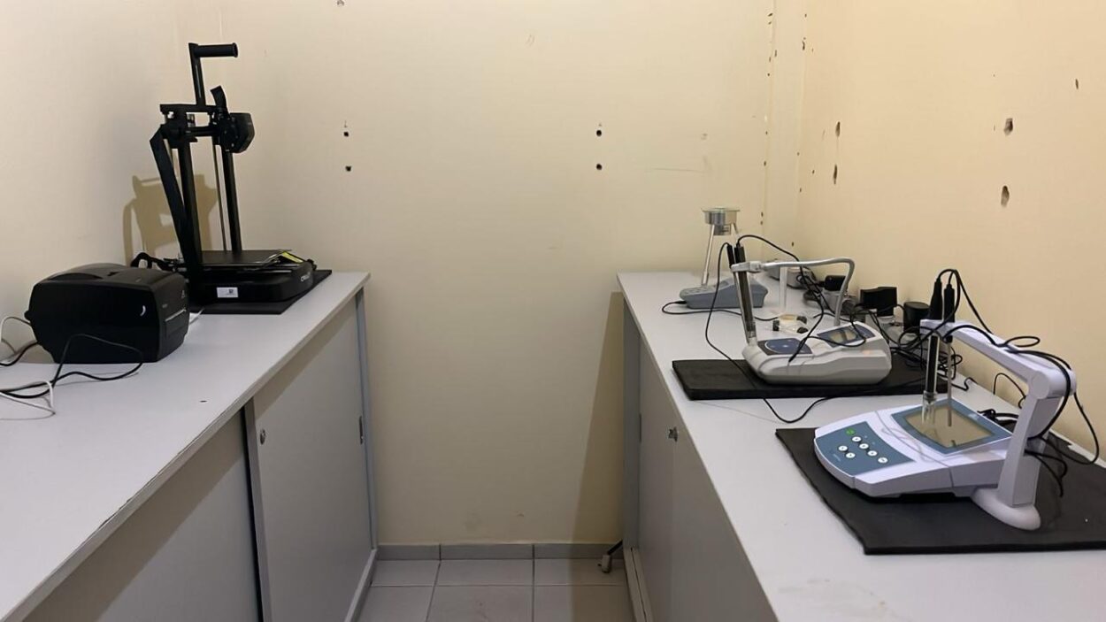 A foto foi tirada em um ambiente fechado, os alunos e sua professora se posicionam lado a lado para a foto, olham diretamente para a câmera. Todos estão usando uniforme da escola, com exceção da profª que está usando calça e jaqueta jeans. Ao fundo, os banners dos trabalhos apresentados.