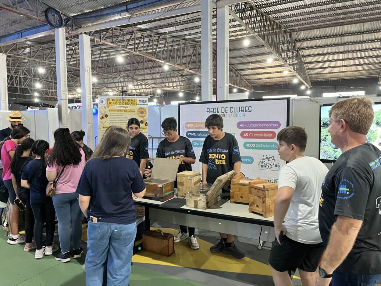 Grupo de jovens e adultos reunidos em um estande de feira dentro de um pavilhão amplo, na Expo Umuarama 2026. Eles observam e manuseiam caixas de madeira sobre uma mesa, nas quais estão armazenadas as colméias de abelhas sem ferrão, enquanto os alunos clubistas explicam o conteúdo. Ao fundo, há painéis informativos do Paraná Faz Ciência e iluminação industrial no teto.