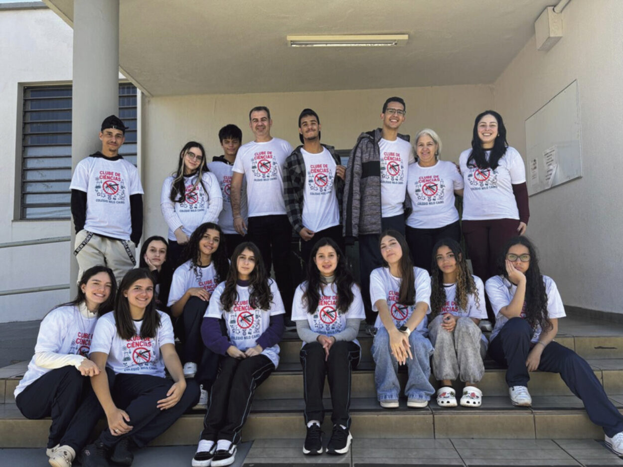 Um grupo de dezessete pessoas, entre adolescentes e adultos, posa em uma escadaria externa de um prédio claro, sob a luz do dia. Eles estão organizados em duas fileiras: alguns em pé nos degraus mais altos e outros sentados nos degraus da frente. Todos vestem camisetas brancas com um símbolo de proibição sobre o desenho de um mosquito, sugerindo uma campanha de combate à dengue. A maioria sorri para a câmera, transmitindo união e entusiasmo. O ambiente é simples, com paredes claras, uma janela lateral e piso cerâmico. A imagem passa a sensação de trabalho coletivo e engajamento em uma ação educativa ou comunitária.