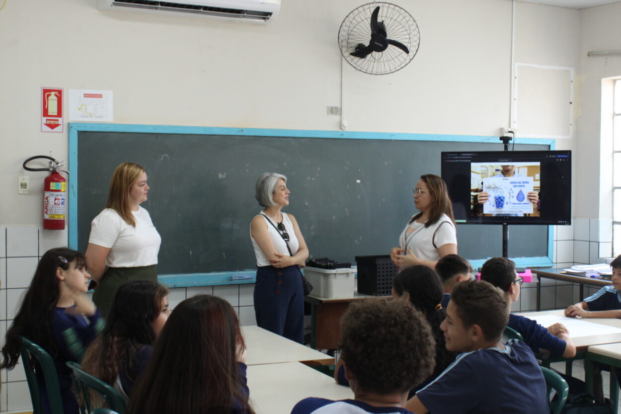 Na foto, uma sala de aula com quadro ao fundo e três pessoas em pé conversando: a professora regente, a articuladora da UEPG e a bolsista responsável pelo clube. À frente, estudantes estão sentados em mesas organizadas em grupos. Há um monitor ligado em uma apresentação de slides sobre o dia da água, tema da atividade realizada pelos clubistas no dia da visita.