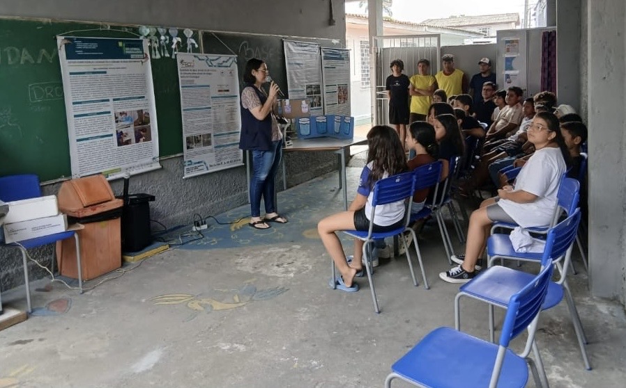Professora Andreia Petruy apresenta conteúdo sobre qualidade da água para alunos sentados em cadeiras, em sala com cartazes científicos ao fundo.