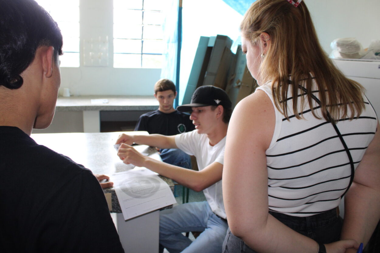 1. A foto mostra um grupo de clubistas reunido em torno de uma mesa em um laboratório, iluminado por luz natural que entra pelas janelas ao fundo. Uma das pessoas está sentada e parece explicar ou mostrar algo em um papel, enquanto as outras observam atentamente, incluindo Fernanda, que está de costas para a câmera. Há materiais sobre a mesa, os estudantes estavam escrevendo seus projetos. Ao fundo, é possível ver uma pia e alguns objetos encostados na parede.