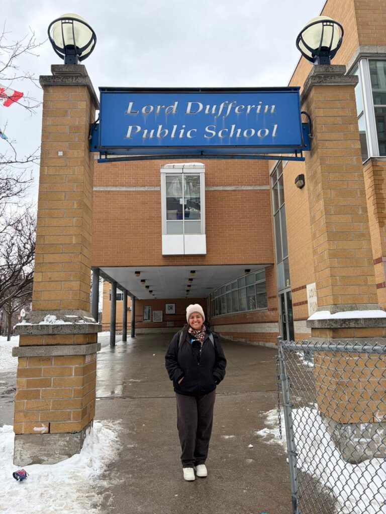 Eloah sorrindo em frente à entrada da Lord Dufferin Public School, em dia frio e com neve no Canadá.