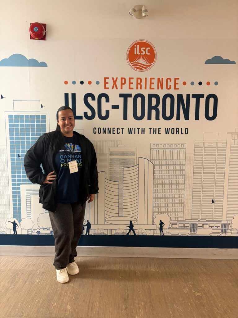 Eloah, sorrindo, posa em frente ao painel “Experience ILSC-Toronto”, com ilustração da cidade ao fundo.