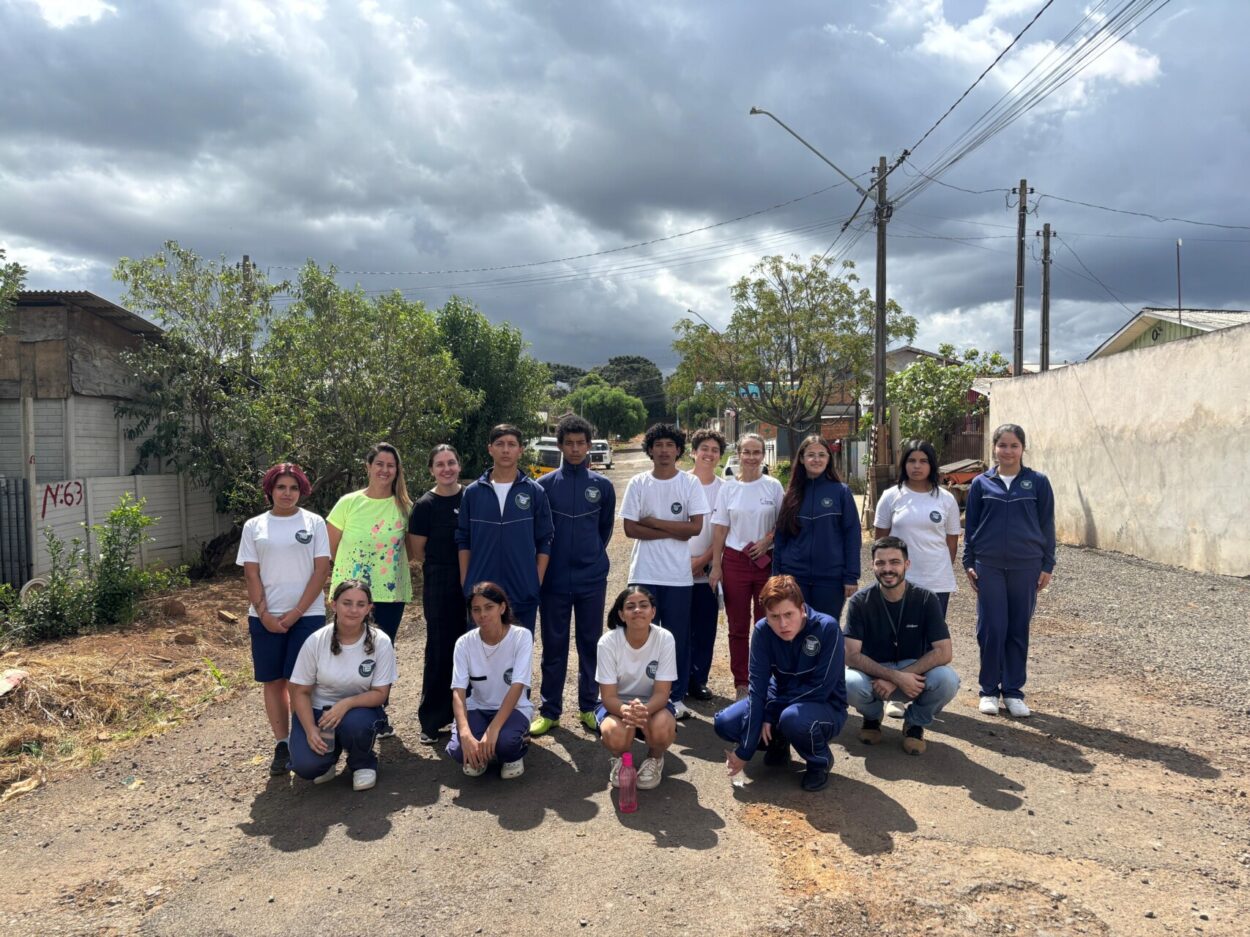 Grupo de cerca de 16 pessoas posando em uma rua de terra em um bairro residencial. A maioria são jovens vestindo uniformes escolares em azul e branco, alguns em pé e outros agachados na frente. Há também alguns adultos junto ao grupo. Ao redor, aparecem casas simples, árvores e postes com fiação elétrica. O céu está parcialmente nublado, com nuvens escuras contrastando com áreas de sol que iluminam o grupo.