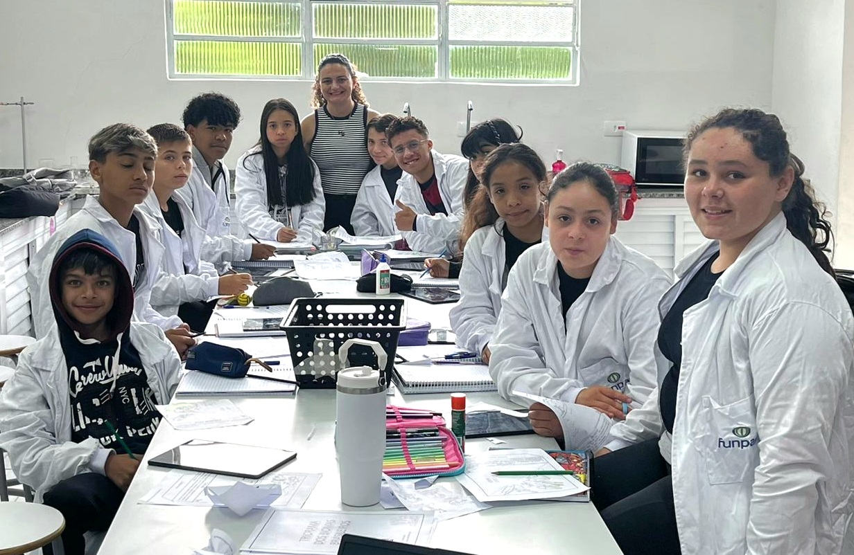 Foto em ambiente escolar mostrando um grupo de adolescentes e uma professora em uma sala clara, sentados ao redor de uma mesa. Todos usam jalecos brancos. Sobre a mesa há cadernos, folhas com atividades, estojo de lápis de cor, cola e garrafa térmica. A professora está ao centro, atrás dos estudantes, e todos olham para a câmera. Ao fundo, há janelas altas e pia, sugerindo um espaço de laboratório ou sala de projetos.