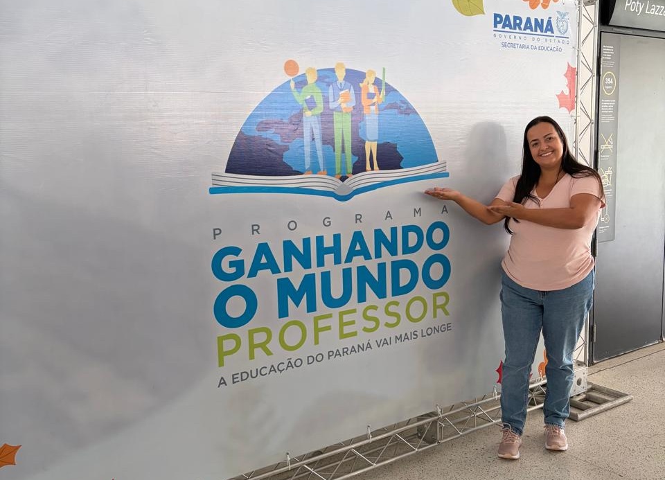 Mulher sorrindo aponta para um painel do programa “Ganhando o Mundo Professor”, do Governo do Paraná, em ambiente interno.