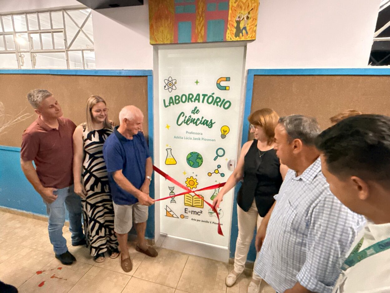 Na foto, um grupo de pessoas, entre professores, autoridades e membros da comunidade escolar estão em frente a entrada do laboratório de ciências durante sua inauguração. As pessoas aplaudem e demonstram alegria. A porta do espaço é decorada com desenhos em ícones representando ciência e pesquisa, como uma lupa, um livro, um DNA, etc, a arte é de Jamille V. Piovesan. Também é possível observar escrito na porta: Laboratório de Ciências Professora Adélia Lúcia Janik Piovesan. Uma fita vermelha está esticada na frente da porta e está sendo cortada com uma tesoura. O ambiente parece ser o interior de uma escola, com painéis de cortiça nas paredes e uma pintura colorida acima da porta, enquanto pequenos pedaços da fita já aparecem no chão, indicando o momento da abertura oficial do espaço.