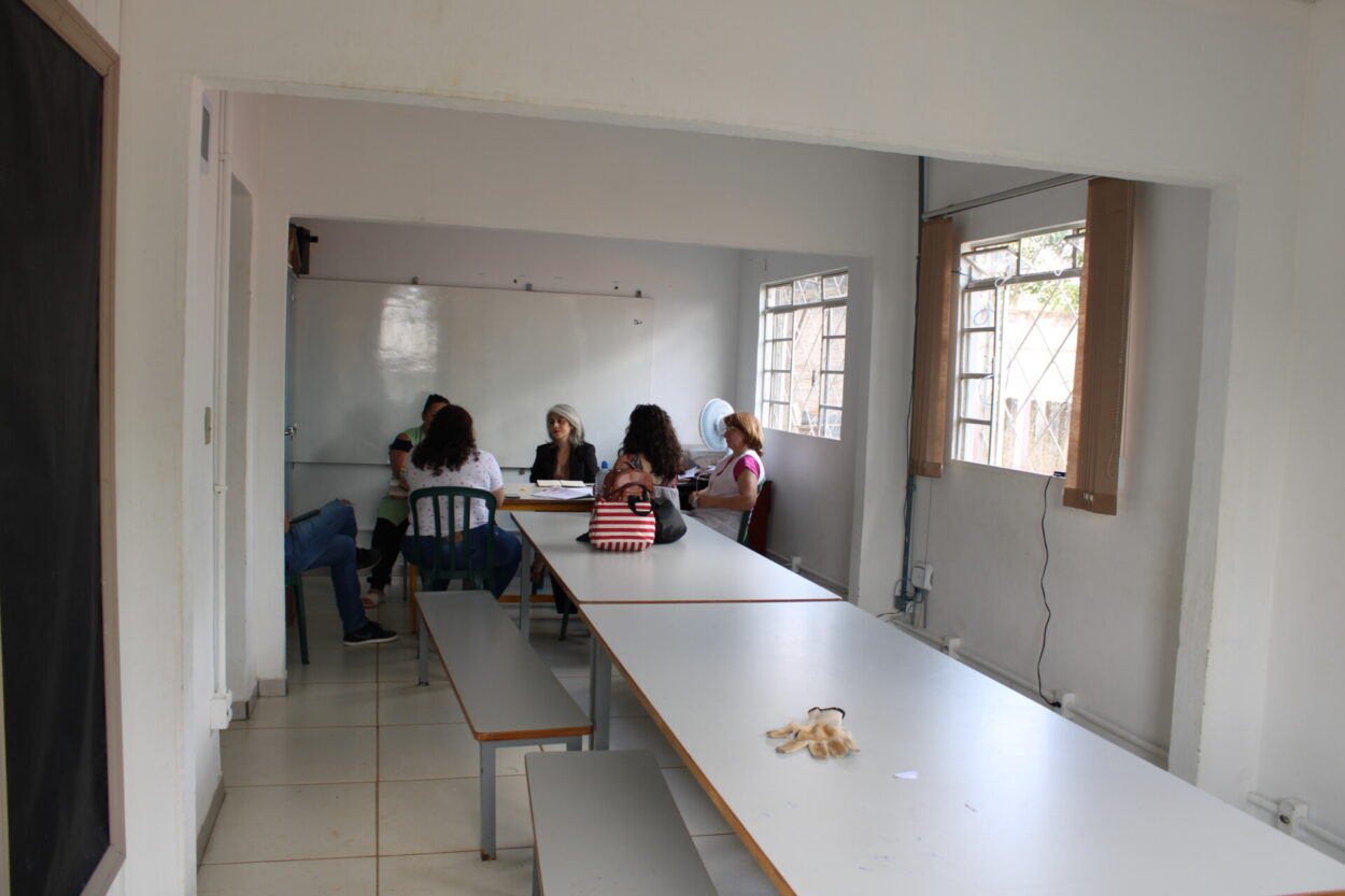 A foto mostra o antigo laboratório. No primeiro plano há mesas longas de madeira com bancos. Ao fundo, um grupo de pessoas está sentado ao redor de uma mesa, conversando e observando alguns papéis. O espaço é iluminado por janelas laterais e possui mobiliário simples, com ventilador e alguns objetos sobre a mesa. 2. Pelo mesmo ângulo da foto anterior, esta foto mostra o novo laboratório de ciências. O espaço possui duas bancadas centrais de mármore com pias e torneiras, além de armários de vidro com vidrarias, equipamentos e modelos didáticos. Ao fundo há uma pequena geladeira e suportes para experimentos, enquanto janelas laterais garantem ventilação e iluminação ao ambiente 3. Em outro ângulo, esta foto também mostra o laboratório novo, desta vez, destaca-se o armário de vidrarias ao fundo, com os equipamentos adquiridos por meio da Rede.
