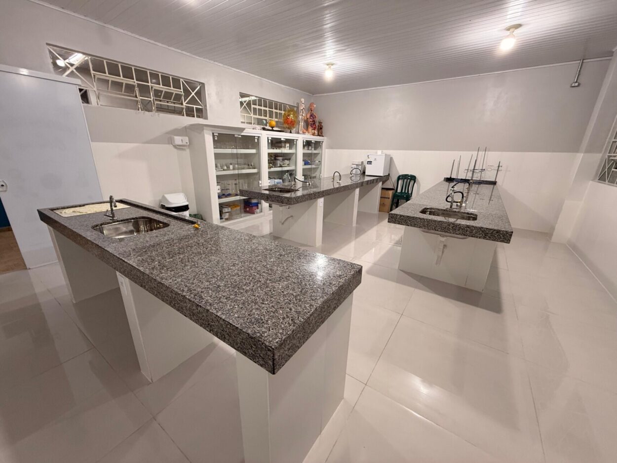 A foto mostra o antigo laboratório. No primeiro plano há mesas longas de madeira com bancos. Ao fundo, um grupo de pessoas está sentado ao redor de uma mesa, conversando e observando alguns papéis. O espaço é iluminado por janelas laterais e possui mobiliário simples, com ventilador e alguns objetos sobre a mesa. 2. Pelo mesmo ângulo da foto anterior, esta foto mostra o novo laboratório de ciências. O espaço possui duas bancadas centrais de mármore com pias e torneiras, além de armários de vidro com vidrarias, equipamentos e modelos didáticos. Ao fundo há uma pequena geladeira e suportes para experimentos, enquanto janelas laterais garantem ventilação e iluminação ao ambiente 3. Em outro ângulo, esta foto também mostra o laboratório novo, desta vez, destaca-se o armário de vidrarias ao fundo, com os equipamentos adquiridos por meio da Rede.