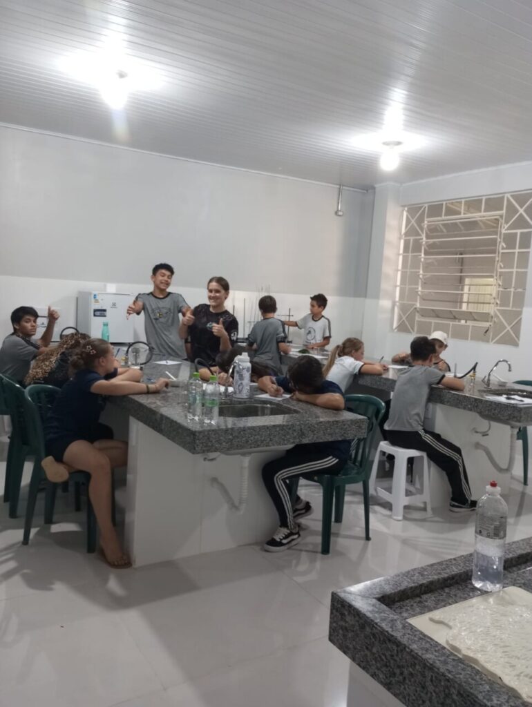 A imagem mostra os clubistas utilizando o novo laboratório de ciências. Eles estão sentados ao redor das bancadas, escrevendo, observando materiais e conversando durante uma atividade. 5. A foto mostra os clubistas realizando uma atividade prática no laboratório. Sobre a bancada estão folhas verdes, recipientes de porcelana, um pilão com pistilo, um béquer com líquido e uma balança digital. Em primeiro plano, uma folha é cortada com uma tesoura e colocada em um recipiente, enquanto outras pessoas ao fundo também manipulam folhas seguindo um roteiro impresso da atividade. 6. A foto mostra um aluno utilizando um microscópio em um laboratório, observando uma lâmina com atenção. O equipamento está sobre uma bancada de pedra, e ao fundo aparecem armários com vidrarias e materiais de laboratório. 7. A foto mostra a professora Rosana ao lado de um microscópio sobre a bancada. Ao fundo aparecem armários com vidrarias e outros materiais científicos, compondo o ambiente de trabalho.