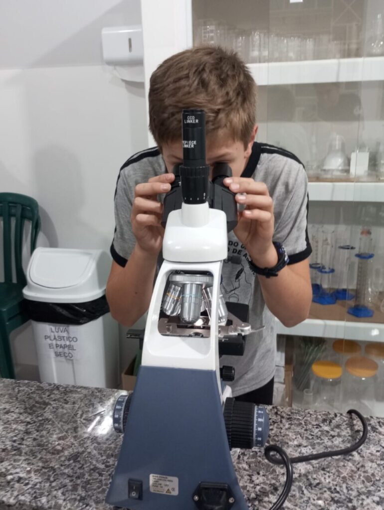 A imagem mostra os clubistas utilizando o novo laboratório de ciências. Eles estão sentados ao redor das bancadas, escrevendo, observando materiais e conversando durante uma atividade. 5. A foto mostra os clubistas realizando uma atividade prática no laboratório. Sobre a bancada estão folhas verdes, recipientes de porcelana, um pilão com pistilo, um béquer com líquido e uma balança digital. Em primeiro plano, uma folha é cortada com uma tesoura e colocada em um recipiente, enquanto outras pessoas ao fundo também manipulam folhas seguindo um roteiro impresso da atividade. 6. A foto mostra um aluno utilizando um microscópio em um laboratório, observando uma lâmina com atenção. O equipamento está sobre uma bancada de pedra, e ao fundo aparecem armários com vidrarias e materiais de laboratório. 7. A foto mostra a professora Rosana ao lado de um microscópio sobre a bancada. Ao fundo aparecem armários com vidrarias e outros materiais científicos, compondo o ambiente de trabalho.