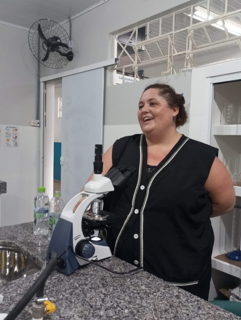 A imagem mostra os clubistas utilizando o novo laboratório de ciências. Eles estão sentados ao redor das bancadas, escrevendo, observando materiais e conversando durante uma atividade. 5. A foto mostra os clubistas realizando uma atividade prática no laboratório. Sobre a bancada estão folhas verdes, recipientes de porcelana, um pilão com pistilo, um béquer com líquido e uma balança digital. Em primeiro plano, uma folha é cortada com uma tesoura e colocada em um recipiente, enquanto outras pessoas ao fundo também manipulam folhas seguindo um roteiro impresso da atividade. 6. A foto mostra um aluno utilizando um microscópio em um laboratório, observando uma lâmina com atenção. O equipamento está sobre uma bancada de pedra, e ao fundo aparecem armários com vidrarias e materiais de laboratório. 7. A foto mostra a professora Rosana ao lado de um microscópio sobre a bancada. Ao fundo aparecem armários com vidrarias e outros materiais científicos, compondo o ambiente de trabalho.