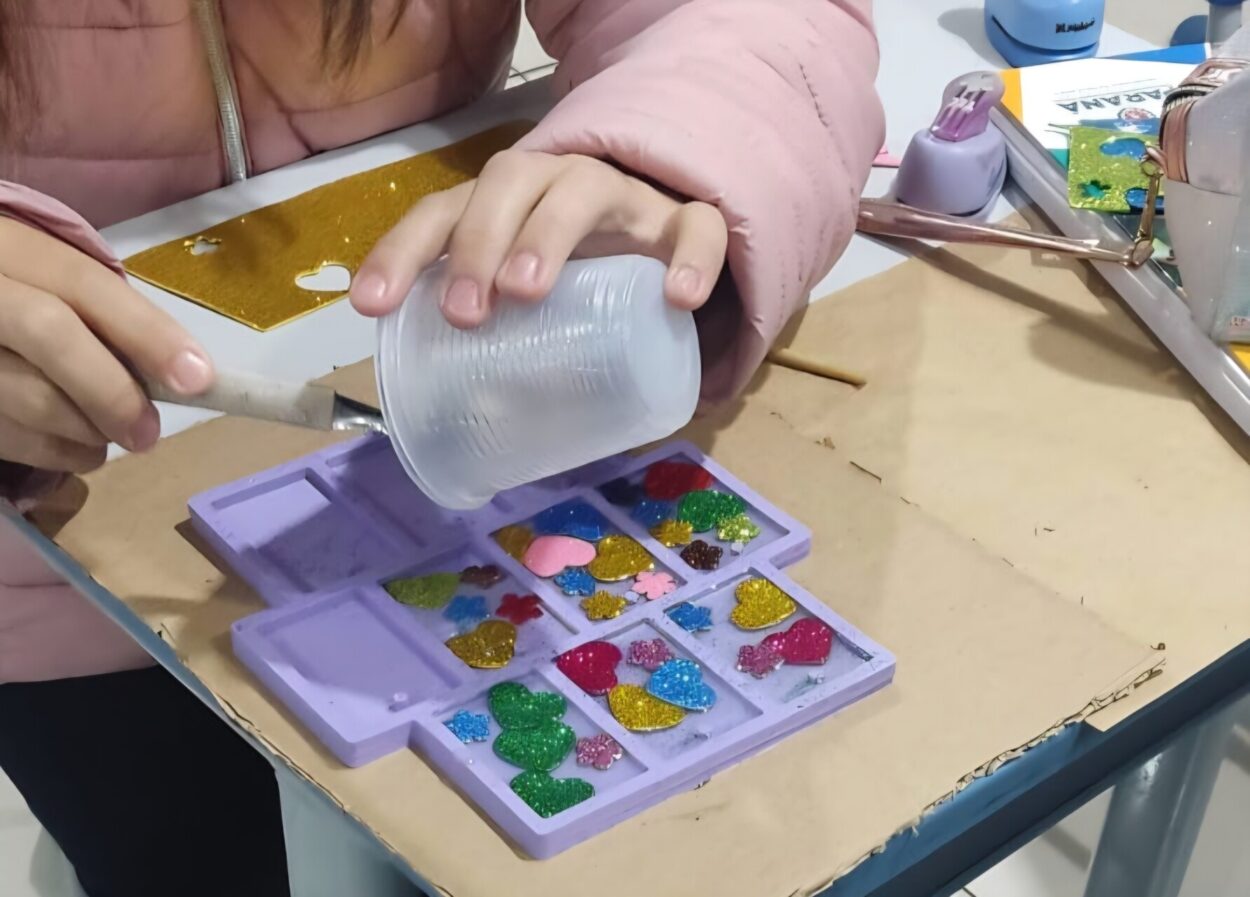 A imagem mostra uma atividade manual ou artesanal sendo realizada sobre uma mesa. No centro da imagem, uma pessoa usa uma jaqueta acolchoada rosa e segura um copo plástico transparente inclinado sobre uma forma de silicone roxa com vários compartimentos. Dentro desses compartimentos há peças decorativas coloridas, muitas em formato de corações, feitas com glitter e cores variadas, como dourado, verde, azul, vermelho e rosa. O copo parece estar sendo usado para despejar líquido, possivelmente resina ou outro material utilizado para moldagem. À esquerda, outra mão segura um utensílio semelhante a uma espátula, auxiliando no trabalho. Ao redor da mesa há diversos materiais de artesanato, como um furador de papel, tesoura, papéis decorativos e outros objetos de trabalho manual.