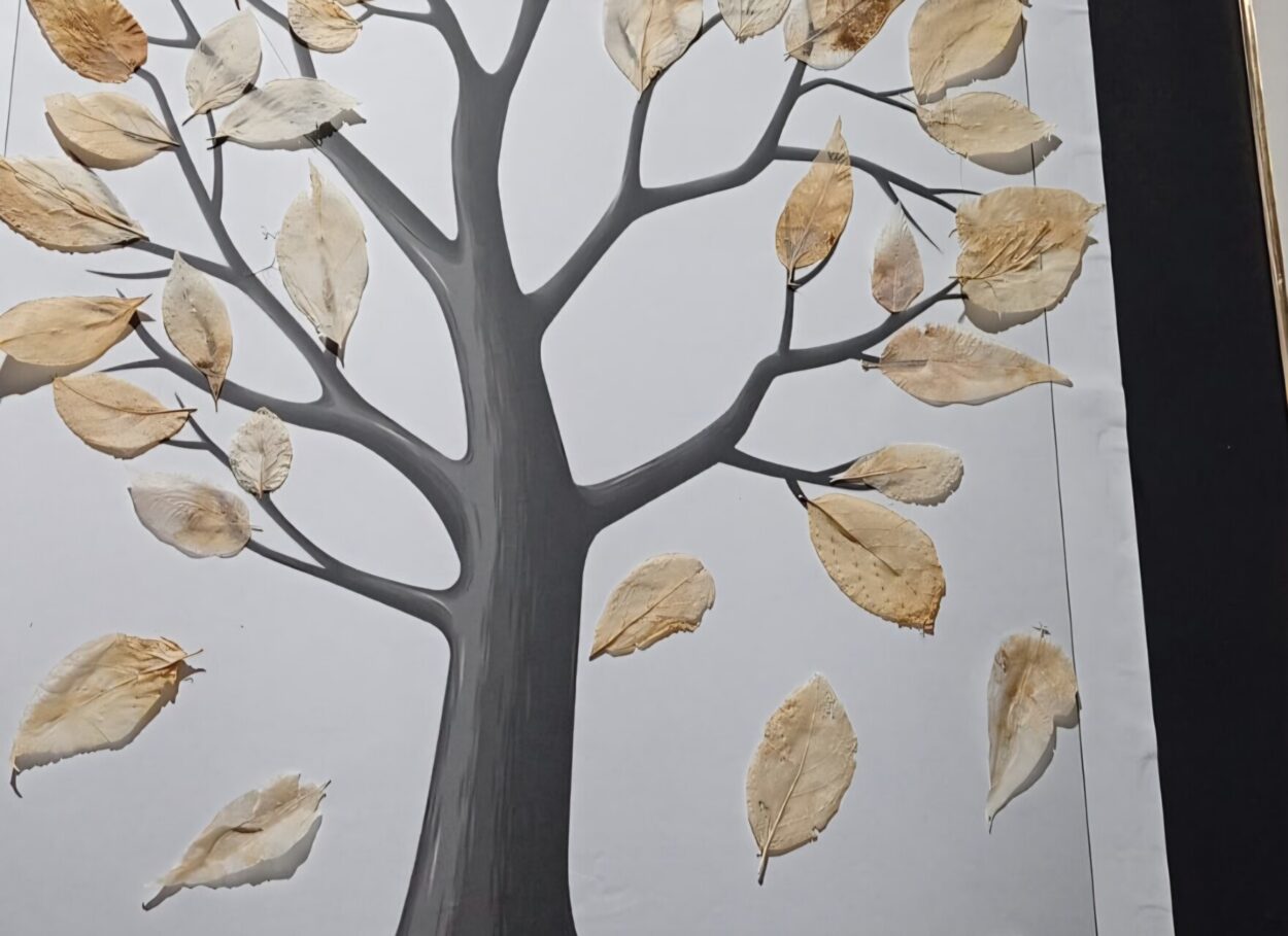 A imagem mostra um painel artístico de uma árvore sobre um fundo branco. O tronco e os galhos da árvore são desenhados ou pintados em tom de cinza. Sobre os galhos estão coladas folhas secas reais, de cor bege ou amarelada. As folhas parecem estar secas e prensadas, algumas com marcas naturais e pequenas rasuras. A composição lembra um trabalho escolar ou artístico de botânica, misturando desenho com elementos naturais. O painel parece estar sobre uma superfície escura, visível nas bordas da imagem.