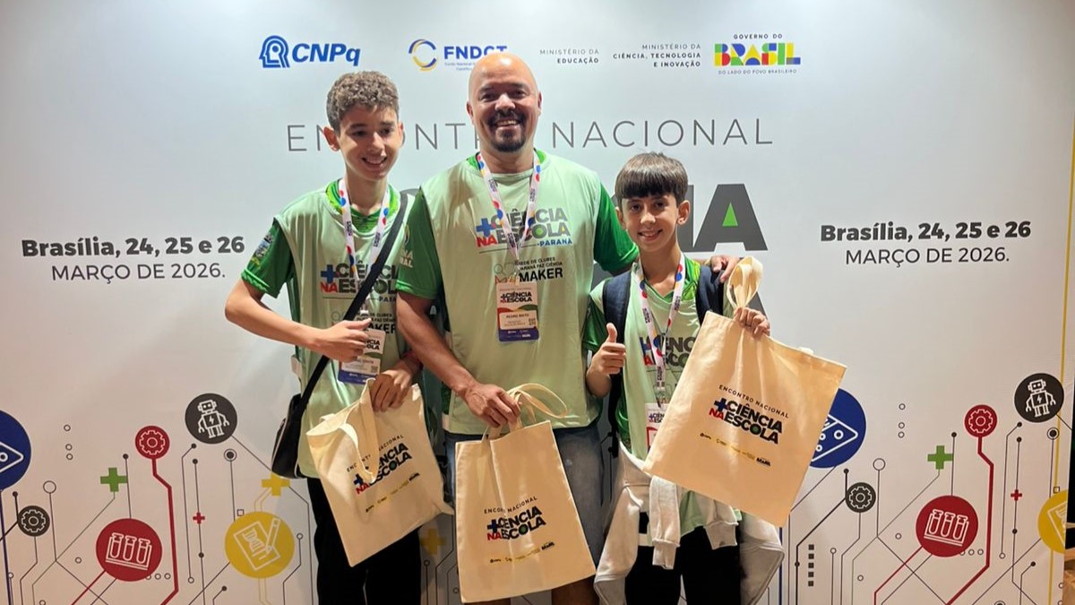 Uma foto em close de três pessoas sorrindo e posando para a câmera em frente a um painel branco de eventos. No centro está o professor Pedro Paulo, um homem de pele morena, careca e de barba, usando óculos de sol pendurados na gola de uma camiseta verde-clara do evento. Ele segura uma sacola de tecido ecobag branca. À sua esquerda está o aluno Rafael Costa, um jovem mais alto usando óculos de grau, camiseta verde do evento e calça escura, também segurando uma ecobag e uma mochila preta. À direita do professor está o aluno Victor Gabriel, o mais jovem da dupla, com cabelos escuros e camiseta verde, segurando sua ecobag branca. As três sacolas têm o logo colorido "Mais Ciência na Escola" impresso. O painel atrás deles é branco com logos de parceiros (CNPq, Governo Federal) e o texto "Encontro Nacional Mais Ciência na Escola - Brasília, Março de 2026". O chão é de piso claro e a iluminação é de spots de luz de um centro de convenções.