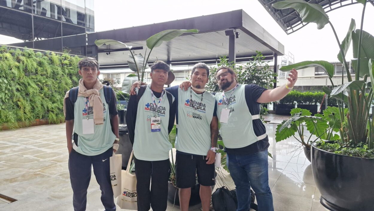 Quatro homens jovens estão lado a lado, posando para uma foto em um espaço moderno com plantas ao redor. Eles usam camisetas claras iguais, com crachás pendurados no pescoço, indicando participação em um evento. Dois deles estão com mochilas. O homem à direita estende o braço, como se estivesse tirando uma selfie ou convidando alguém para a foto. Ao fundo, há um ambiente com cobertura, paredes de vidro e bastante vegetação, sugerindo um local de evento ou instituição.