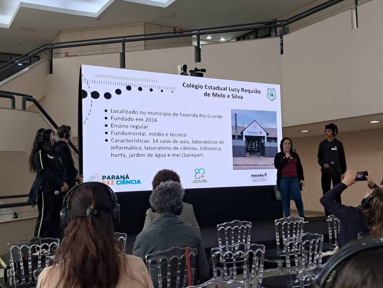 As imagens mostram os integrantes do clube Liga Científica durante sua apresentação. No palco, estudantes e a professora compartilham o projeto com o público, apoiados por um painel de slides. Três fotos da galeria destacam os resultados da pesquisa diretamente da tela de apresentação.
