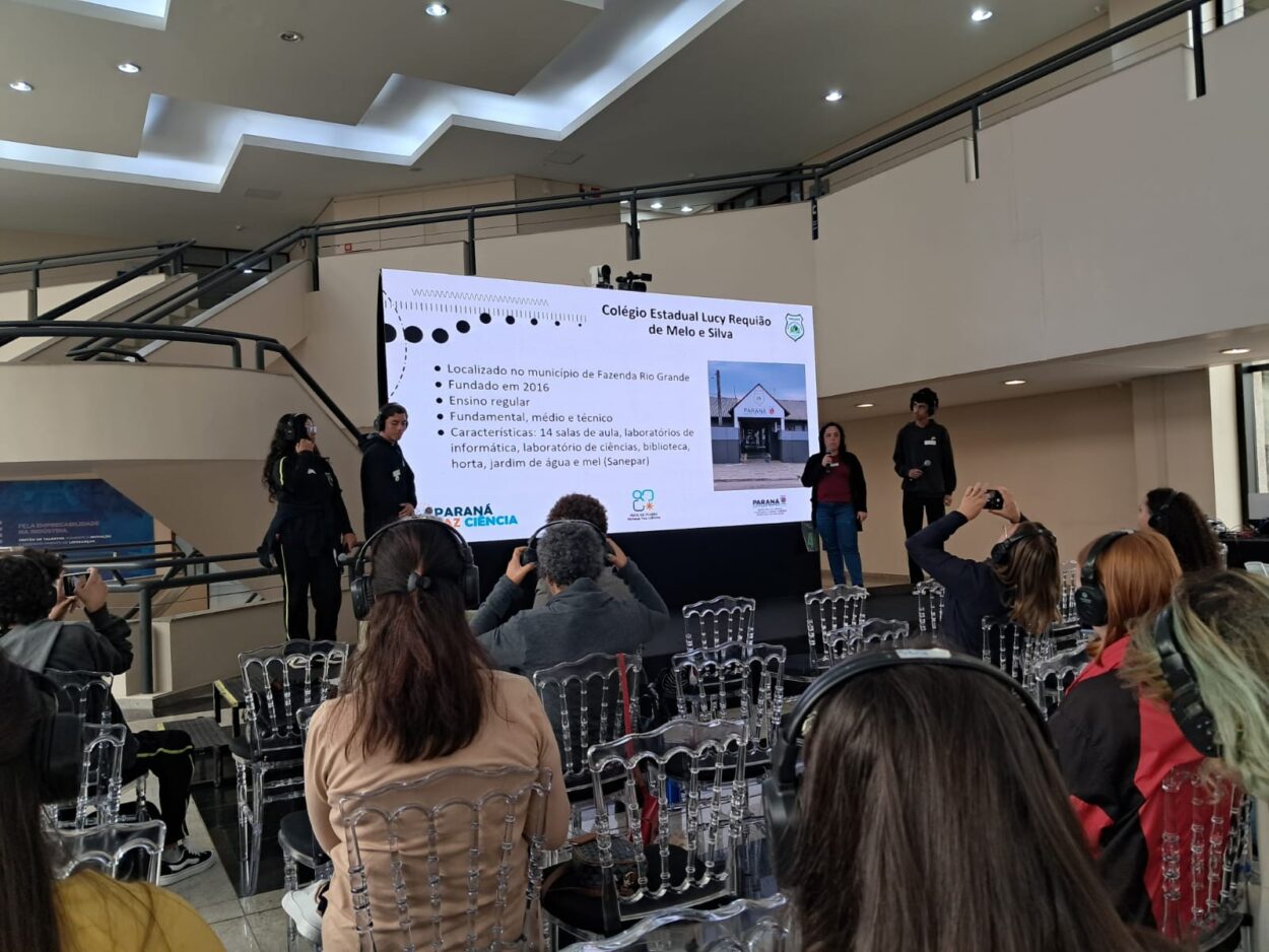 As imagens mostram os integrantes do clube Liga Científica durante sua apresentação. No palco, estudantes e a professora compartilham o projeto com o público, apoiados por um painel de slides. Três fotos da galeria destacam os resultados da pesquisa diretamente da tela de apresentação.