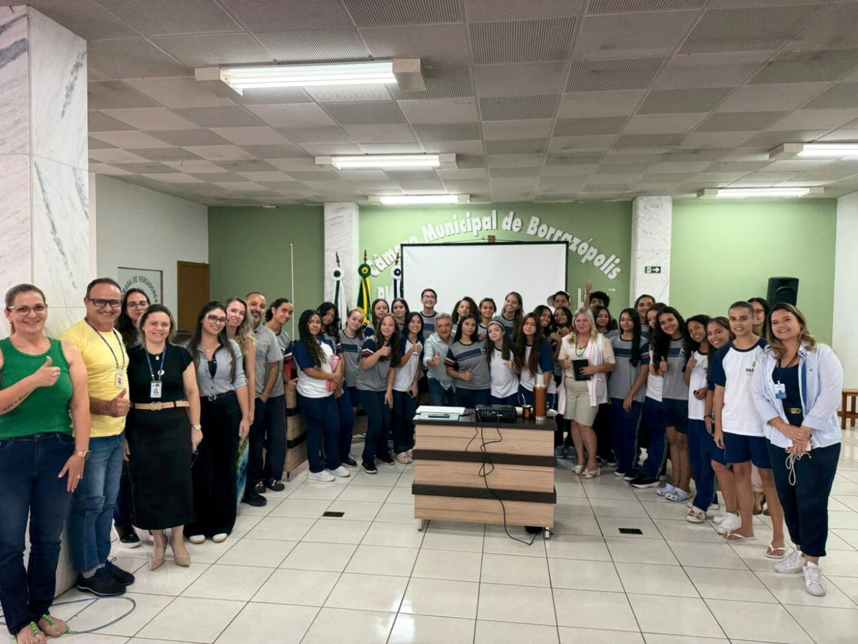 Um grupo grande de estudantes e adultos está reunido dentro de um auditório ou sala de eventos. Eles estão organizados em um semicírculo ao redor de uma mesa com computador e projetor, posicionada no centro da sala. Ao fundo, há uma tela de projeção e uma parede verde com a inscrição “Câmara Municipal de Borrazópolis”. A maioria dos estudantes veste uniforme escolar em tons de azul, branco e cinza. Alguns adultos estão ao lado do grupo, usando roupas casuais ou de trabalho. Várias pessoas sorriem e algumas fazem gesto de positivo com o polegar, demonstrando entusiasmo. O espaço é iluminado por lâmpadas fluorescentes no teto quadriculado, e o piso é de cerâmica clara. A imagem transmite a ideia de um encontro educativo ou apresentação coletiva em um espaço público.