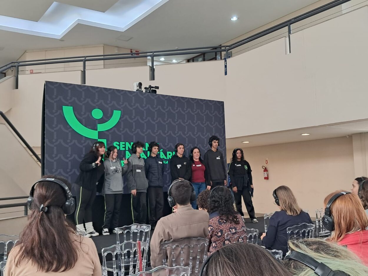Fotografia em ambiente interno, em um espaço de eventos. No centro, um grupo de cerca de oito estudantes e uma professora está sobre um pequeno palco, posando para uma foto. Eles usam roupas casuais e uniformes escuros, e estão lado a lado, alguns sorrindo. Ao fundo, há um grande painel preto com um símbolo verde estilizado e a palavra “Semana Araucária”, indicando o nome do evento. Na frente do palco, várias pessoas estão sentadas em cadeiras, de costas para a câmera, assistindo à apresentação. Algumas usam fones de ouvido, sugerindo tradução simultânea ou acessibilidade sonora. O ambiente é bem iluminado, com paredes claras e um mezanino ao fundo. A cena transmite um momento de apresentação ou participação em evento educacional ou científico.