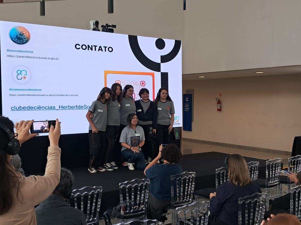 As imagens mostram os integrantes do clube de ciências Mentes em Ação durante sua apresentação. No palco, cinco estudantes e professora compartilham o projeto com o público, apoiados por um painel de slides. Três fotos da galeria destacam os resultados da pesquisa diretamente da tela de apresentação.