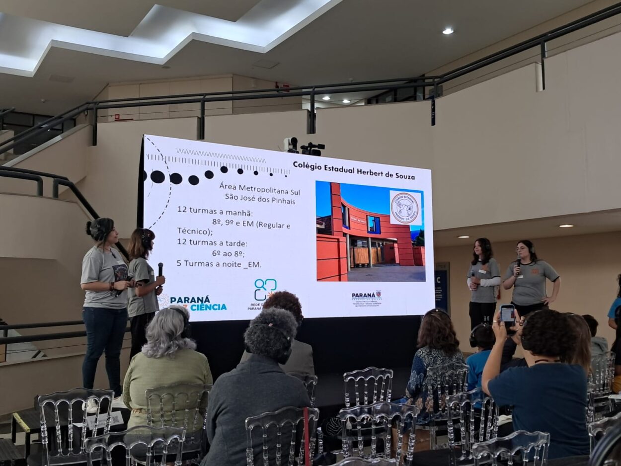 As imagens mostram os integrantes do clube de ciências Mentes em Ação durante sua apresentação. No palco, cinco estudantes e professora compartilham o projeto com o público, apoiados por um painel de slides. Três fotos da galeria destacam os resultados da pesquisa diretamente da tela de apresentação.