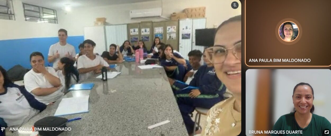 Grupo de estudantes e um professor em sala de aula, organizados em fileiras de carteiras. Um dos alunos segura um caderno aberto. Na tela, a bolsista Bruna aparece em vídeo chamada, acompanhando a atividade