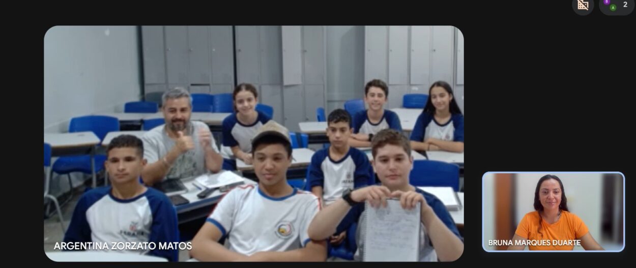 Estudantes reunidos em sala de aula, posando juntos e sorrindo para a câmera. Ao lado, em uma janela de videochamada, a bolsista pedagógica Bruna também aparece sorrindo, indicando participação remota no encontro do clube