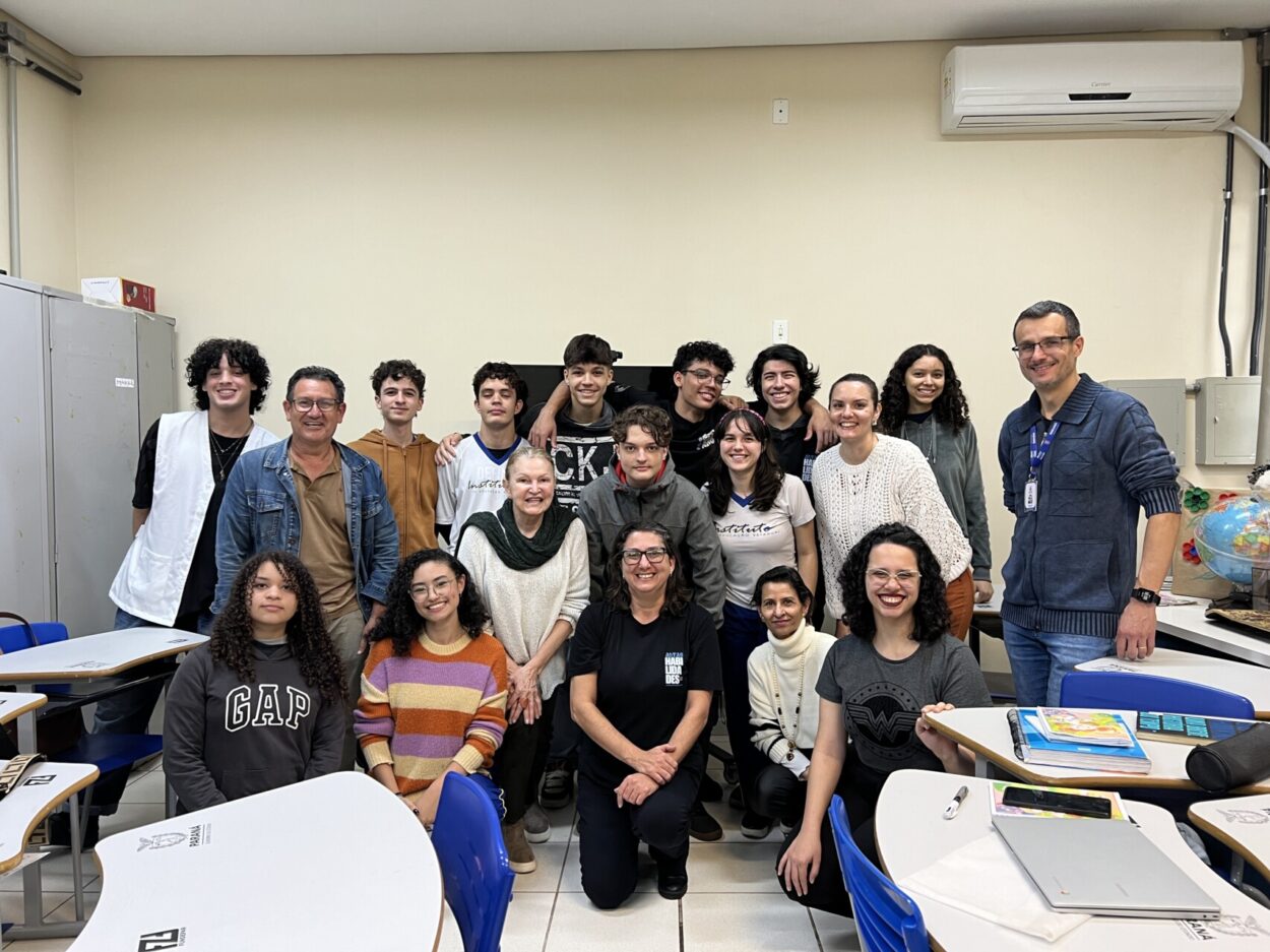 A foto é do Clube de Ciências ‘Cienteens” do Colégio Instituto Estadual de Educação de Maringá. A imagem mostra um grupo grande de pessoas dentro de uma sala de aula. São adolescentes e adultos. Eles estão todos posando para a foto juntos, muitos sorrindo. A sala tem carteiras escolares brancas com detalhes em madeira clara e assentos azuis. No fundo, há armários metálicos, televisão/monitor em cima de um suporte, uma lousa clara e um ar-condicionado instalado na parede superior direita. Também é possível ver um globo terrestre sobre uma mesa no canto direito, dando a ideia de um ambiente pedagógico. A maior parte do grupo está de pé atrás e no centro da imagem, formando uma fileira, enquanto algumas pessoas estão sentadas ou agachadas na frente.