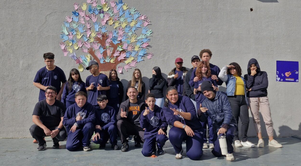 A imagem mostra um grupo grande de adolescentes e alguns adultos reunidos ao ar livre, posando para uma foto. Eles estão organizados em duas fileiras: alguns agachados na frente e outros em pé atrás. A maioria veste uniforme escolar azul-escuro. Ao fundo, há uma parede clara com um desenho de uma árvore feito com vários recortes coloridos em formato de mãos, formando a copa da árvore. À direita, há um pequeno cartaz colorido com desenhos e elementos decorativos. O grupo parece descontraído: muitos estão sorrindo, fazendo gestos com as mãos (como sinal de positivo ou poses informais). A iluminação indica que é dia, com sol forte iluminando a cena.