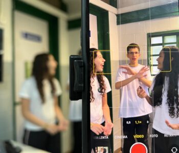 A imagem mostra três jovens em uma sala de aula. Eles estão sendo filmados pela câmera de um celular, cuja tela aparece em destaque. No centro, um rapaz usa óculos e gesticula com as mãos enquanto fala. Ao lado dele, duas moças sorrindo parecem conversar com ele. Todos vestem uniforme do colégio. O fundo tem paredes claras com detalhes em verde e uma janela.