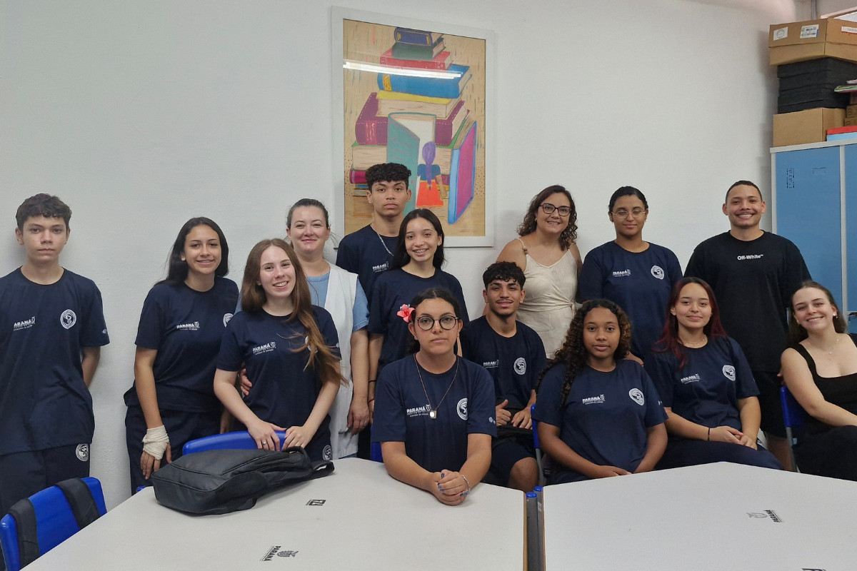 Esta imagem mostra um grupo de estudantes e alguns adultos dentro de uma sala de aula. Eles estão reunidos para uma foto, organizados em duas fileiras: alguns em pé atrás e outros sentados ou apoiados nas mesas à frente. A maioria usa camisetas azul-escuras com um logotipo, sugerindo uniforme escolar. Ao fundo, há uma parede branca com um quadro decorativo que mostra uma pilha de livros coloridos. À direita, aparece um armário azul e algumas caixas empilhadas. As mesas estão à frente do grupo, com mochilas e materiais escolares sobre elas.
