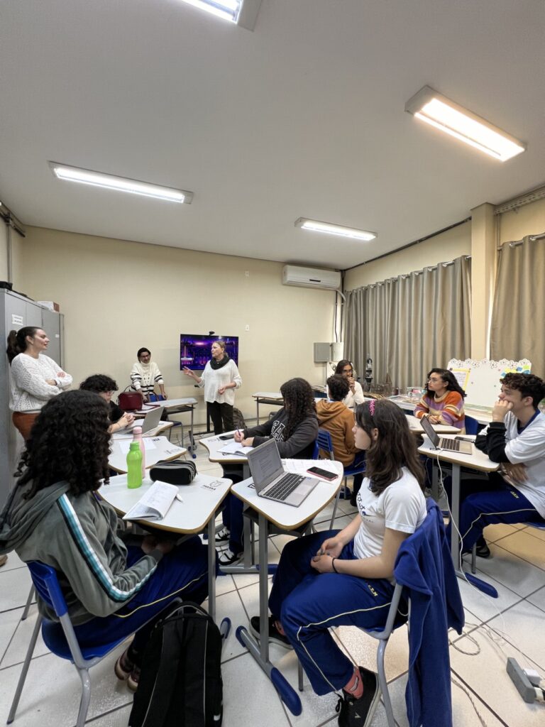 A imagem mostra uma sala de aula com um grupo de estudantes sentados em mesas organizadas em pequenos agrupamentos. As pessoas estão concentradas. Na parte frontal da sala há três pessoas em pé. Ao fundo, há uma TV ligada na parede, mostrando uma imagem colorida. A sala possui iluminação branca de teto, cortinas fechadas do lado direito e um ar-condicionado na parede de trás.