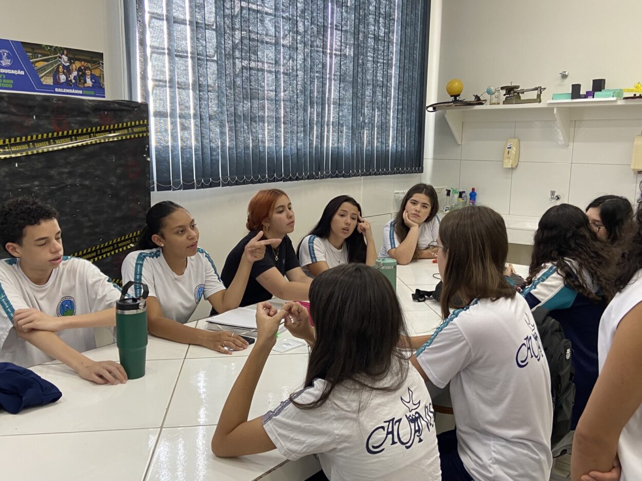 Na foto, nove clubistas do Cabeceira do Rio Iapó estão reunidos conversando, sentados, ao redor de uma mesa. O espaço em que se encontram é o laboratório da escola, nele, uma ampla janela fechada por uma cortina com persianas, uma estante com materiais expositivos, uma pia, um painel e paredes claras. Os estudantes estão com uniformes da escola e parecem focar sua atenção na pauta da conversa.