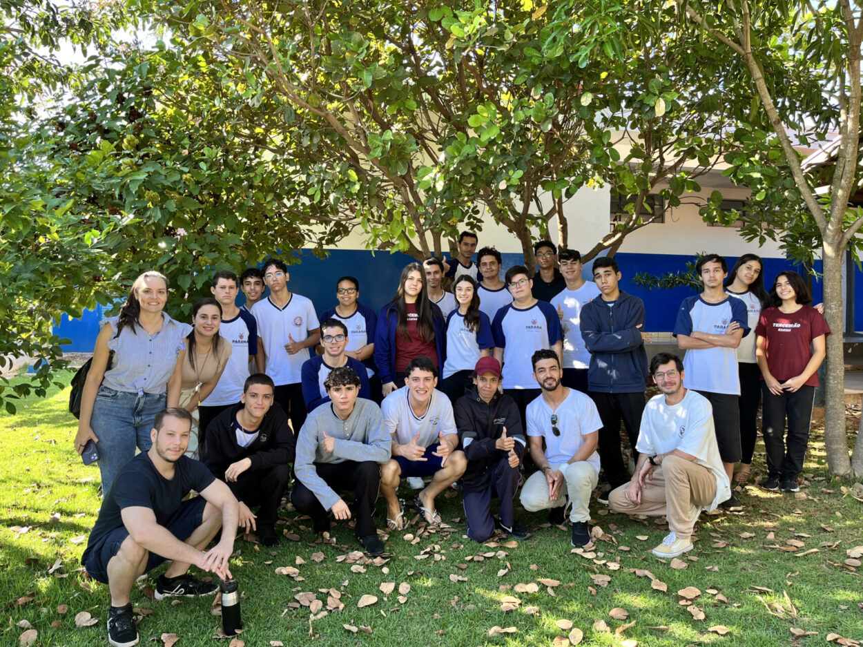 Foto de um grupo grande de pessoas ao ar livre, estudantes e professores. Cerca de 20 jovens e alguns adultos posam juntos em frente a um prédio com parede branca e faixa azul. Eles estão sob árvores com muitas folhas verdes, que fazem sombra sobre o grupo.Na frente, alguns estão agachados ou sentados na grama, enquanto os demais ficam em pé atrás. A maioria veste camisetas claras com detalhes escuros, nas cores do uniforme escolar. Todos estão próximos, alguns sorrindo, em um clima descontraído. No chão há folhas secas espalhadas, e a luz do sol ilumina bem a cena.
