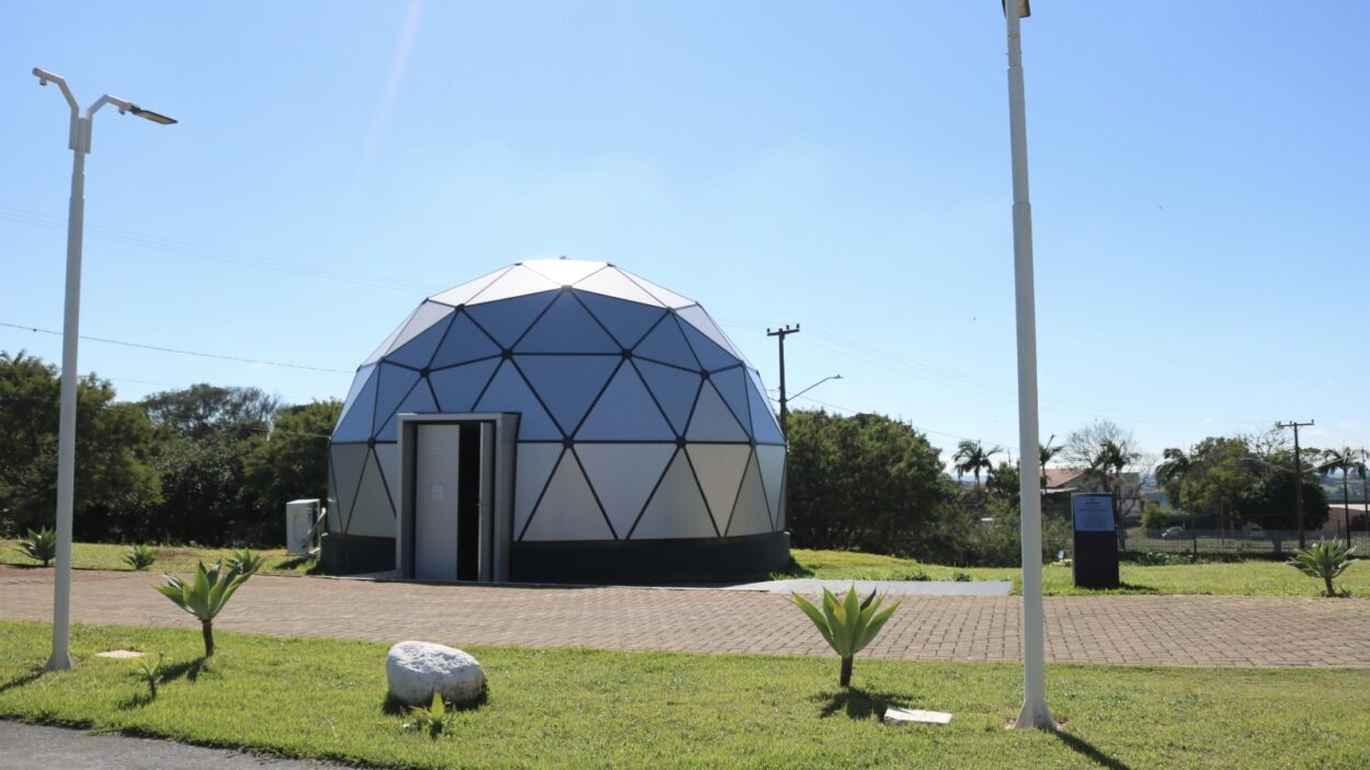 Vista externa do planetário da Universidade Estadual do Norte do Paraná, em Cornélio Procópio. A construção tem formato de cúpula geodésica, com painéis triangulares em tons claros e estrutura escura. A porta de entrada está aberta na parte frontal. O planetário fica em uma área ao ar livre, com gramado bem cuidado, pequenas plantas ornamentais e um caminho de piso intertravado à frente. Há postes de iluminação ao redor e, ao fundo, árvores e algumas construções. O céu está claro e ensolarado, indicando um dia de tempo aberto.
