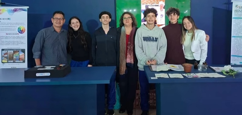 A imagem mostra um grupo de sete pessoas posando em um espaço de exposição científica, durante uma feira de ciências. O grupo é formado por adultos e jovens estudantes. Todos estão lado a lado, sorrindo para a câmera, atrás de duas mesas de apresentação. No centro há uma mulher usando óculos e um cachecol, aparentando ser professora e orientadora do grupo. Há dois banners científicos posicionados nas laterais: à esquerda, um banner sobre um projeto relacionado ao uso de cores e pigmentos. À direita, outro banner com textos científicos, imagens e gráficos sobre experiências interdisciplinares envolvendo artes e ciências. Sobre as mesas há documentos e folhas impressas, recipientes e objetos de experimento, pequenos materiais de demonstração, uma planta/folhagem exposta, e uma caixa de apresentação.